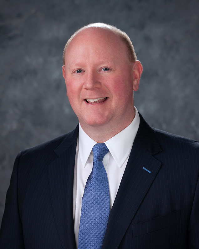 Man in navy suit and blue tie smiling for a professional headshot