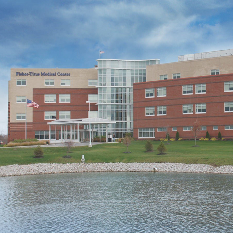 Exterior view of Fisher-Titus Medical Center, a multi-story brick building beside a pond with an American flag