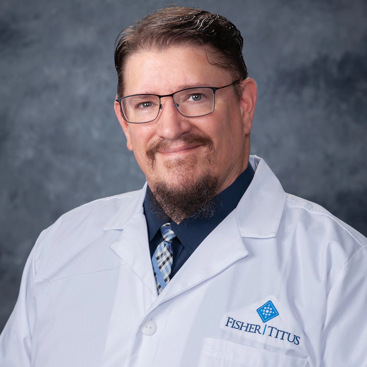 Smiling male Fisher-Titus physician wearing glasses, a dark suit, and white lab coat