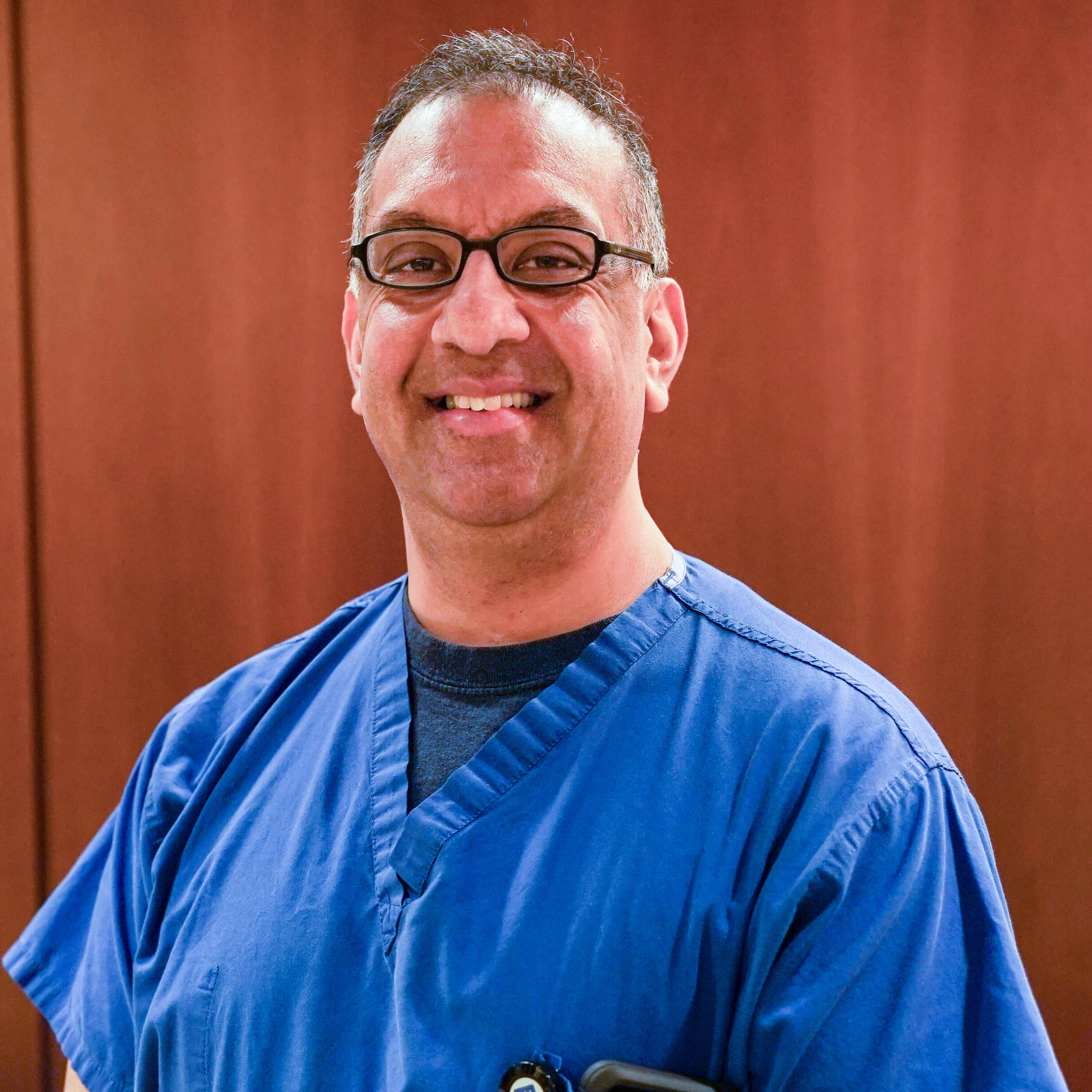 Smiling male healthcare professional wearing blue scrubs and glasses, posed for a professional staff portrait at Fisher-Titus.